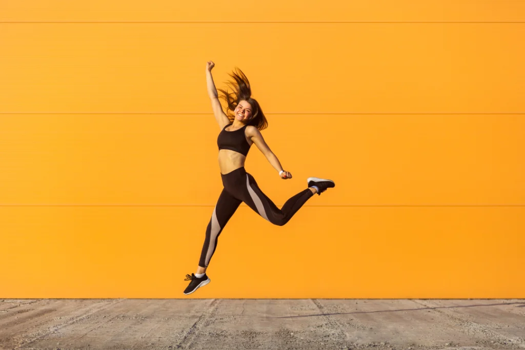 A fit and happy woman jumping in front of an orange wall, showcasing the potential fitness results of CoolSculpting.