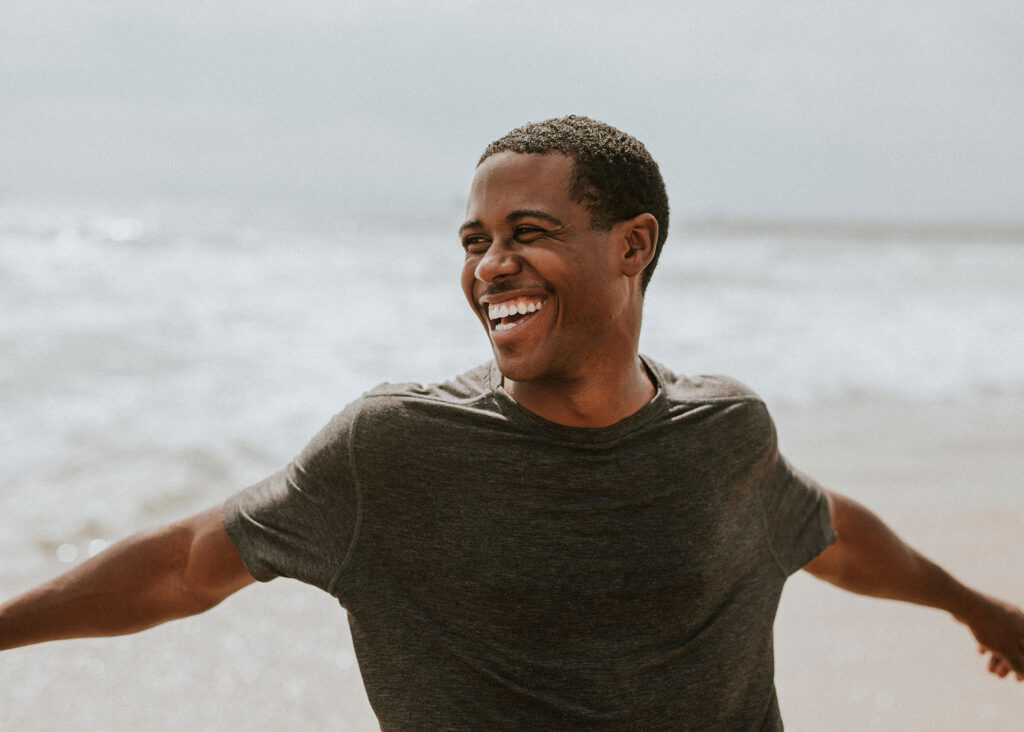 man in a gray shirt standing near the ocean with arms open and smiling broadly