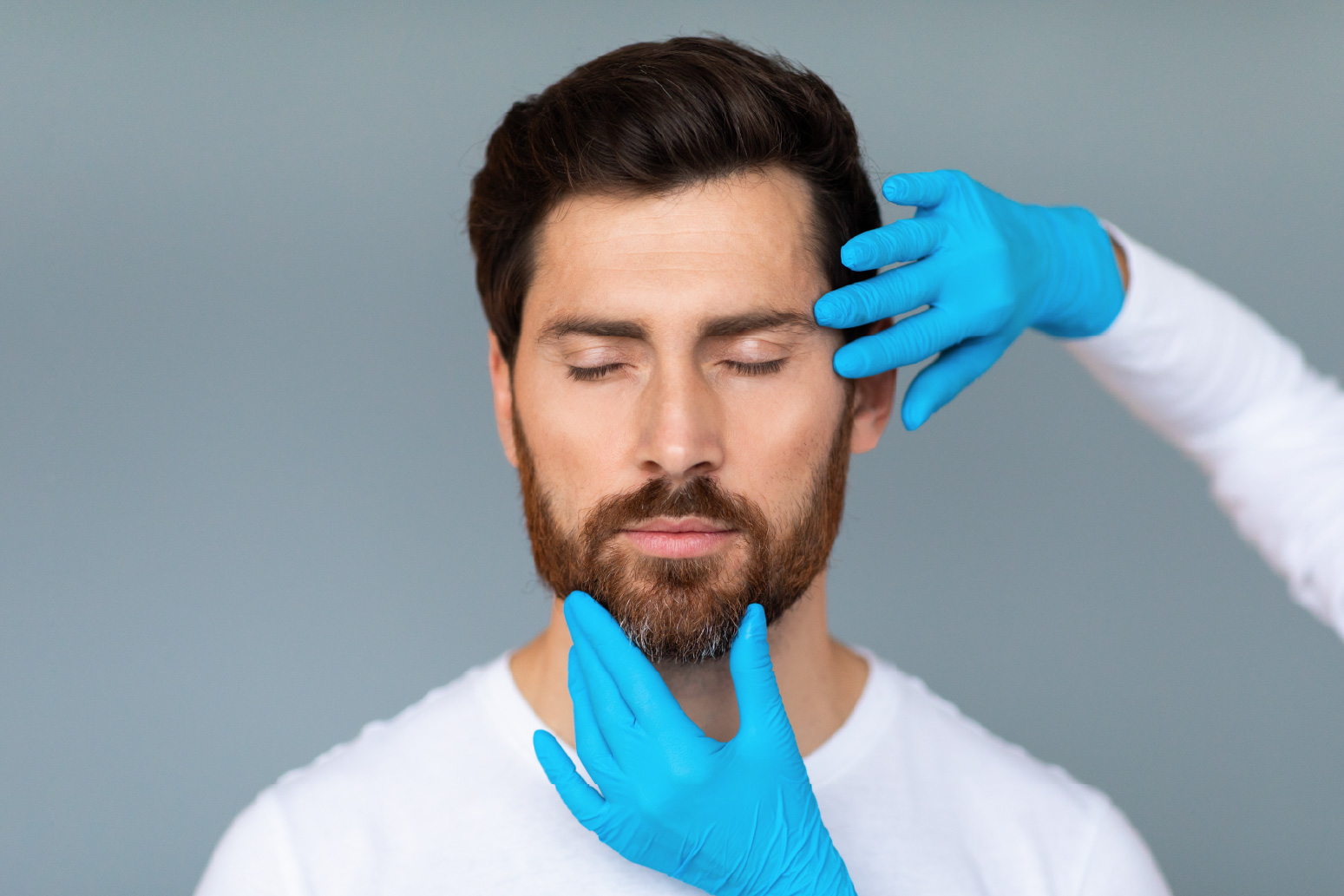 Man with closed eyes being examined by gloved hands touching his face and chin against a gray background.