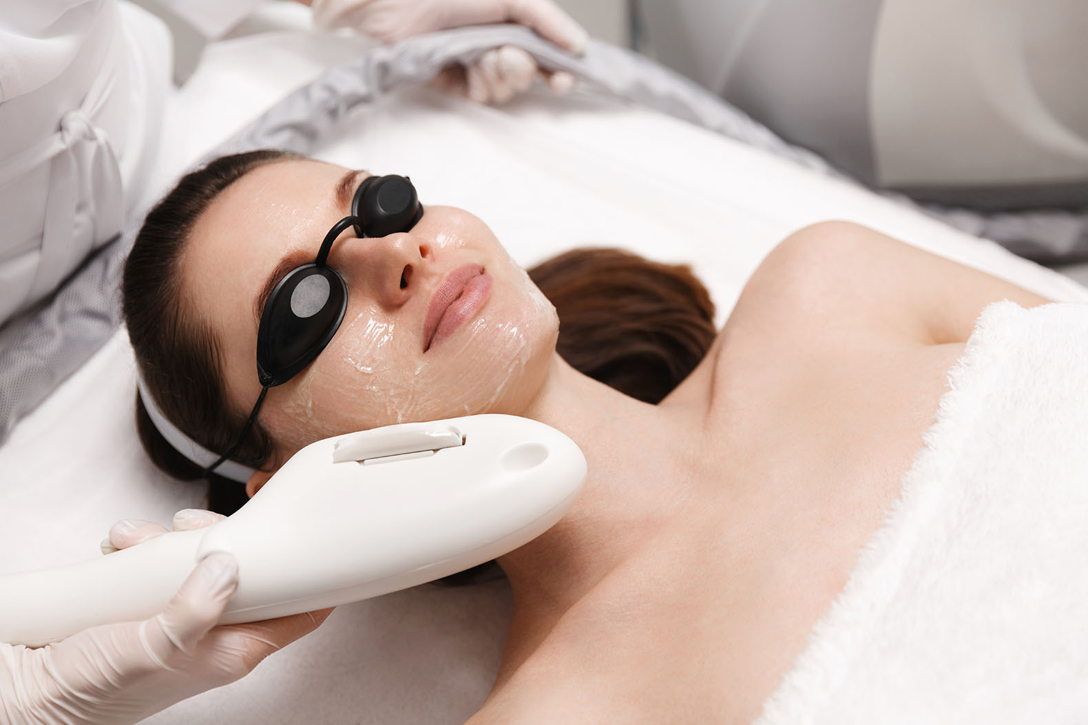 Person lying with protective goggles while a gloved hand holds a light-based device near the face, which is coated with a gel.