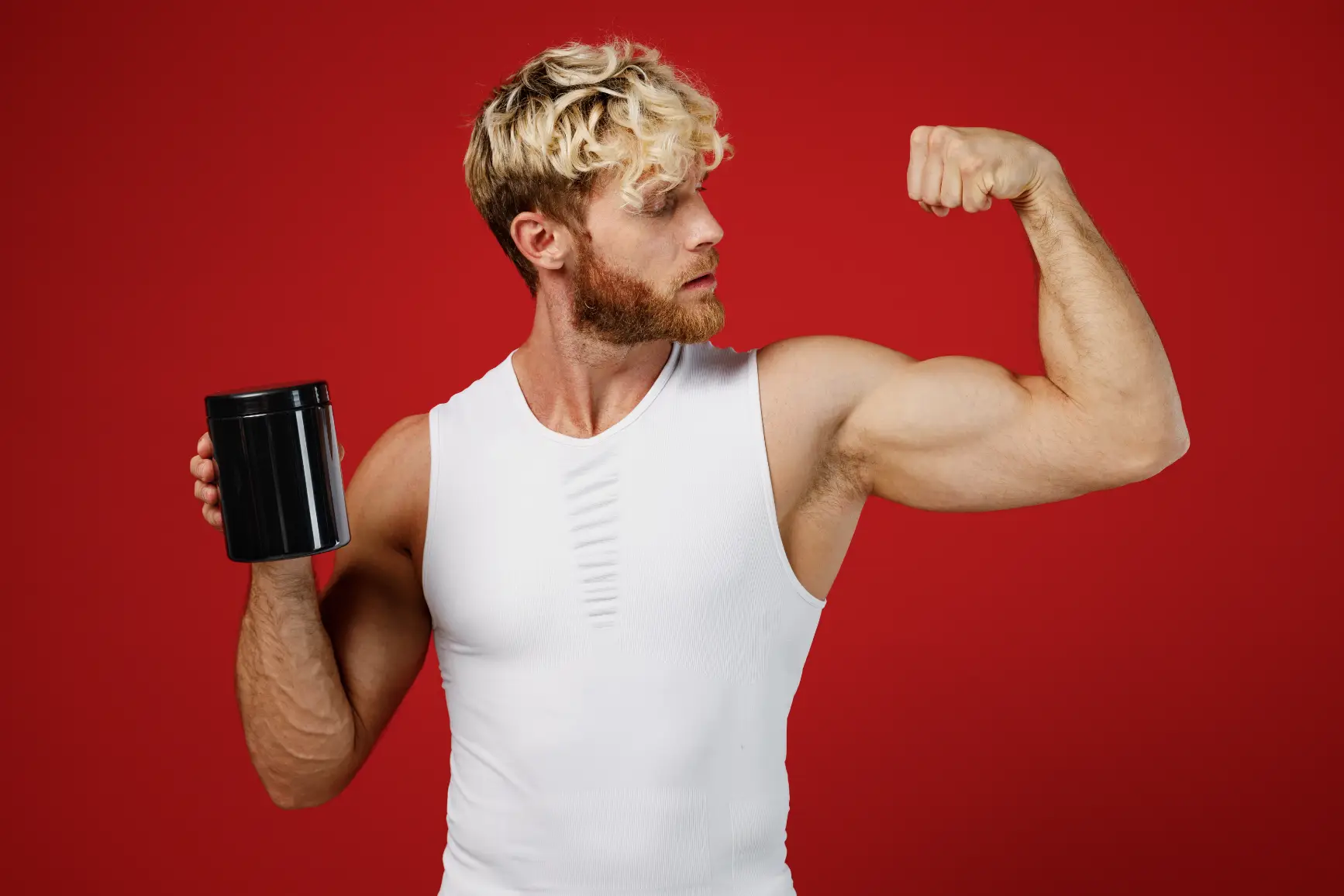 Muscular man flexing arm while holding supplement container against red background