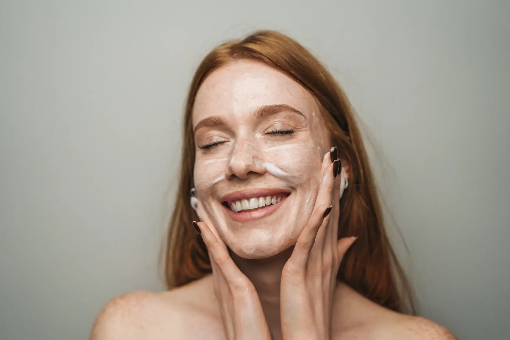 Smiling woman applying foaming cleanser to her face with eyes closed