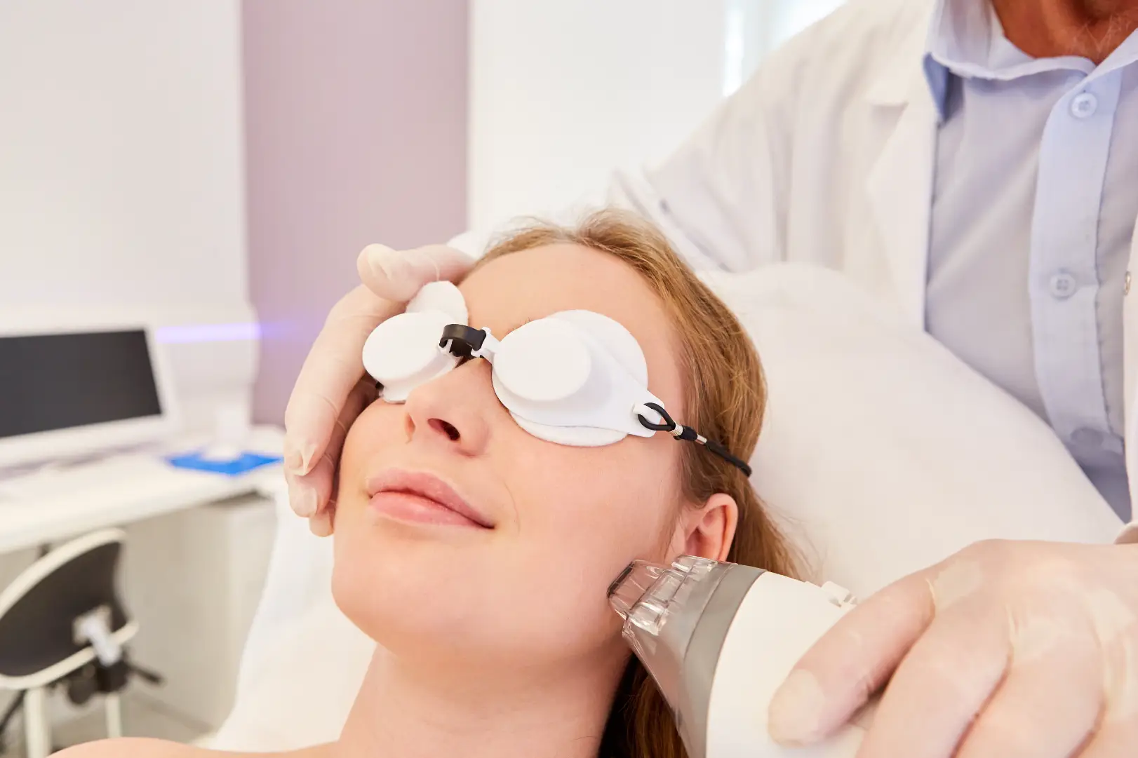 Woman wearing protective eye covers while a practitioner holds a treatment device against her cheek.