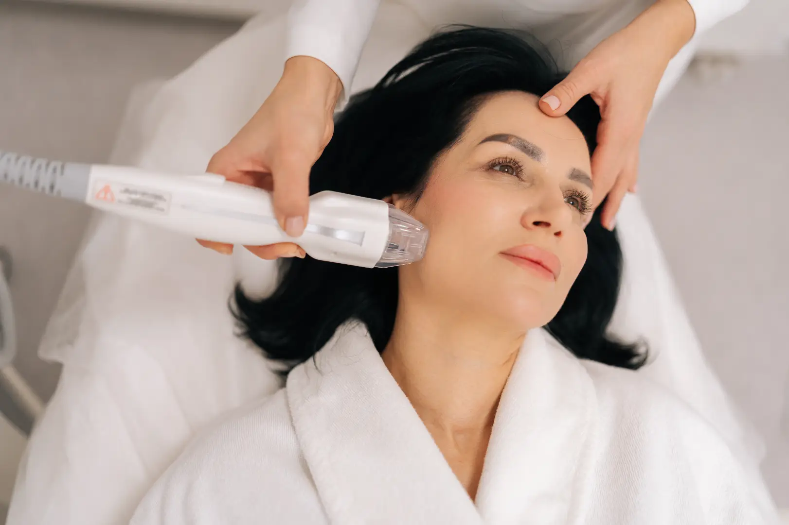 Practitioner holds a handheld device against a woman's cheek while she reclines on a treatment bed.