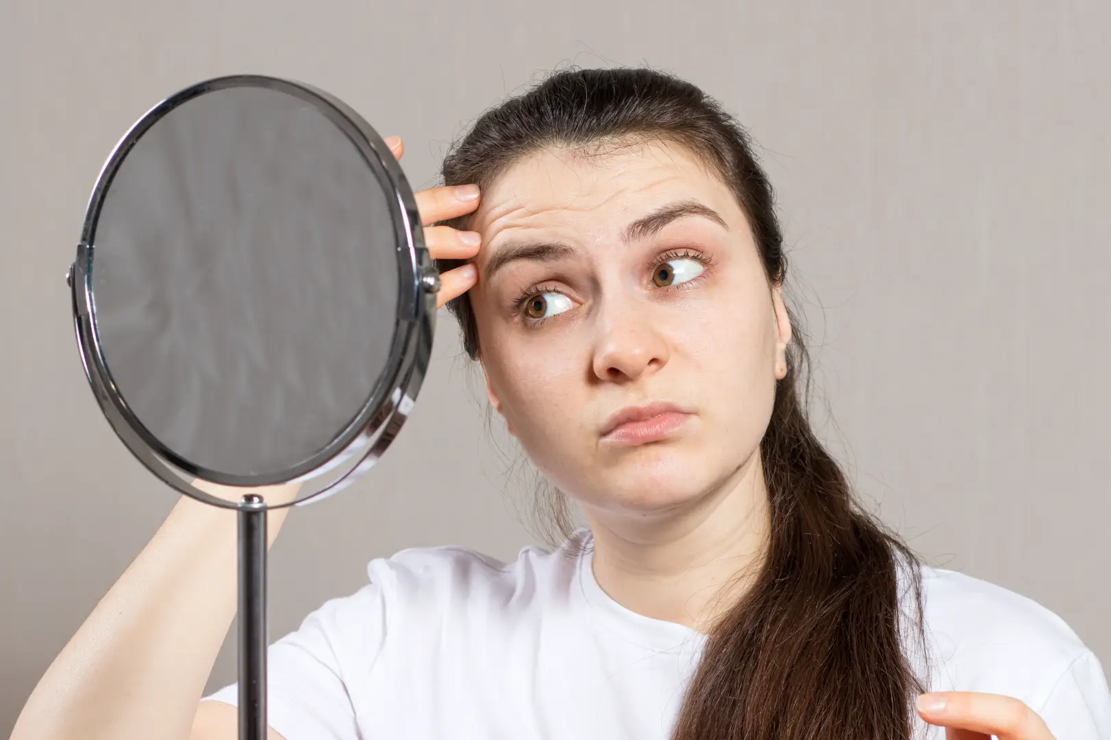 Woman looks into a tabletop mirror while touching the skin near her forehead and temple.